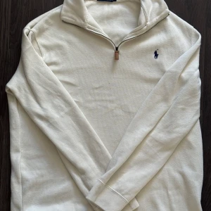 Vit tröja med dragkedja från Polo Ralph Lauren - Säljer en stilren vit tröja från Polo Ralph Lauren med dragkedja och broderad logga på bröstet. Tröjan har långa ärmar och en klassisk krage. Perfekt för en avslappnad och elegant look.