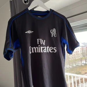  Chelsea fotbollströja retro 2003 - Säljer en svart Chelsea fotbollströja från Umbro med Fly Emirates som sponsor. Tröjan har korta ärmar och blå detaljer längs sidorna och ärmarna. Lite dekekter på sponsorn men annars bra skick 
