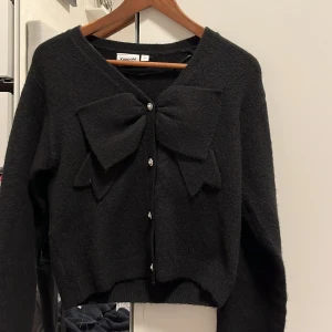 Svart kofta med rosett från Kappahl - Snygg svart kofta med stora rosetter framtill och knappar. Perfekt för en stilren look. Långärmad och mjukt material som ger en bekväm passform.