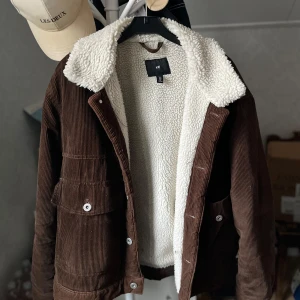 Brun manchesterjacka med teddyfoder - Säljer en snygg brun manchesterjacka från Hm med mjukt vitt teddyfoder. Jackan har knappar framtill och två stora fickor med lock. Perfekt för kyligare dagar med sin varma och stiliga design.