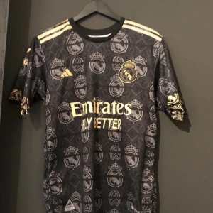 Svart Real Madrid fotbollströja från Adidas - Säljer en svart Real Madrid fotbollströja från Adidas med guldiga detaljer och klubbens emblem. Tröjan har korta ärmar och ett mönster med klubbens logga. Texten 'VINI JR.' och nummer 7 pryder baksidan. Perfekt för fans av laget!