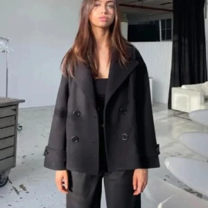 Svart kappa med knappar - Snygg svart kappa med dubbelknäppning och breda slag. Jackan har en stilren design med långa ärmar och en lös passform. Perfekt för en chic look.