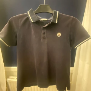 Svart pikétröja från Moncler - Säljer en stilren svart pikétröja från Moncler med klassisk krage och knappar. Tröjan har korta ärmar och en broderad logotyp på bröstet. Perfekt för en avslappnad men ändå elegant look.Snygg att ha till Sommaren.Tröjan i bild är inte tvättad men kommer såklart ny tvättad🍾🔥