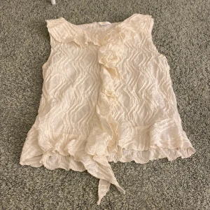 Beige blus från Gina Tricot - Säljer en elegant beige blus från Gina Tricot med volangdetaljer framtill. Blusen är ärmlös och har en fin struktur i tyget som ger en vacker look! Denna är helt ny och köpte för 229!