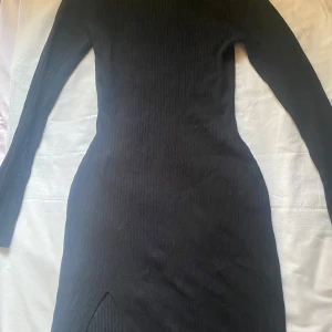 Svart ribbad klänning - Snygg svart ribbad klänning med lång ärm och en subtil slits framtill. Perfekt för en stilren look. Klänningen har en tight passform som framhäver figuren- smal vid midjan