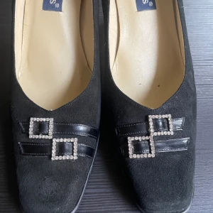 Svarta pumps med dekorativa detaljer - Eleganta svarta pumps i mocka med fyrkantig tå och klack. Skorna har dekorativa detaljer i form av glittriga fyrkanter och svarta remmar på ovansidan. Perfekta för en stilren look.