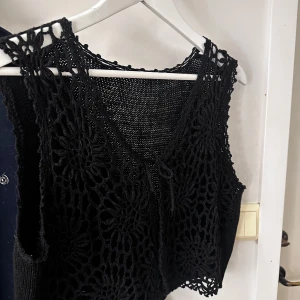 Svart virkad väst - Snygg svart virkad väst med blommönster. Toppen är ärmlös och har en v-ringad design. Perfekt för att lägga till en bohemisk touch till din outfit. Fler bilder går att få.