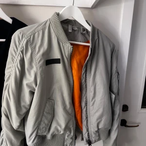 Grå bomberjacka från H&M Divided - Snygg grå bomberjacka från H&M Divided med orange foder. Jackan har dragkedja framtill och ribbade muddar vid ärmslut och nederkant. Perfekt för en cool och avslappnad stil.
