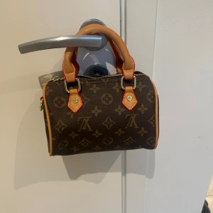 Brun handväska med monogram - Snygg brun handväska med klassiskt monogrammönster och kontrasterande handtag i beige och rött. Väskan har en dragkedja upptill och guldfärgade detaljer. Perfekt för att ge en touch av lyx till din outfit. Har en liten liten defekt på under sidan av väskskan men inget man tänker på (Det tillkommer även ett längre band) 