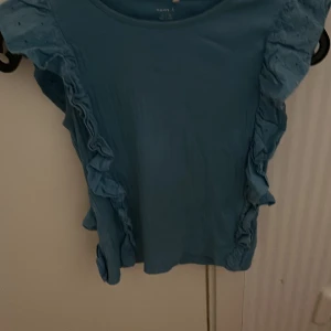Blå topp från Name It - Säljer en söt blå topp från Name It med volangdetaljer längs sidorna. Perfekt för en stilren och lekfull look. Toppen har en rund halsringning och är tillverkad i ett mjukt material.