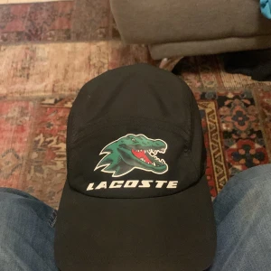 Svart keps från Lacoste - Snygg svart keps från Lacoste med det klassiska krokodilmärket framtill. Kepsen har en justerbar rem baktill för perfekt passform. Perfekt för sportiga aktiviteter eller en casual look.