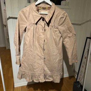 Beige kappa från Odd Molly - Säljer en beige kappa från Odd Molly med volangdetaljer vid nederkanten och knappar framtill. Jackan har långa ärmar och en unik design med en färgglad insida. Perfekt för att ge din outfit en stilfull touch.