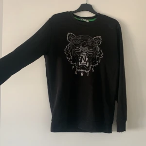 Svart tröja från Kenzo med tigerdesign - Säljer en svart tröja från Kenzo med en cool tigerdesign på framsidan. Tröjan har långa ärmar och en rund halsringning. Perfekt för dig som vill ha en stilren men ändå iögonfallande look.