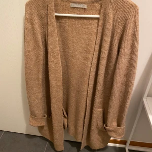 Beige stickad kofta från By Malene Birger - Mysig beige stickad kofta från By Malene Birger. Perfekt för kyliga dagar med sina långa ärmar och öppna design. Koftan har praktiska fickor framtill och ribbade muddar vid ärmsluten. Ett stilrent plagg som passar till många tillfällen.