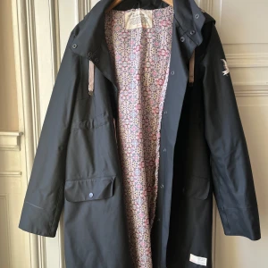 Svart regnjacka odd Molly  - Snygg svart regnjacka från Odd Molly med ett färgglatt blommigt foder. Jackan har långa ärmar och praktiska fickor framtill. Den är utrustad med knappar och dragsko i huvan för extra skydd mot regn. Perfekt för regniga dagar! Använd få tal gånger, lite slitage i tyget, skicka meddelande för mer info, mått och bilder, hund finns i hemmet :) storlek 2 = M/L upplever den stor i storleken 