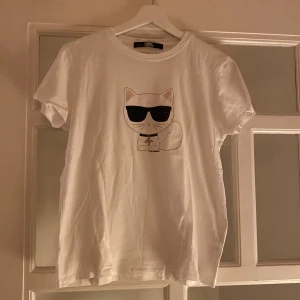 Vit t-shirt från Karl Lagerfeld - Snygg vit t-shirt från Karl Lagerfeld med ett coolt kattmotiv som bär solglasögon. Saknas någon sten på solglasögonen men inget som syns. Använd 2 gånger.
