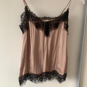 Beige linne med spetsdetaljer - Säljer ett elegant beige linne med svarta spetsdetaljer. Linnet har tunna axelband och vacker spets längs halsringningen och nederkanten. Perfekt för en stilren look.