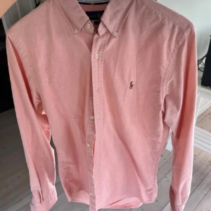 Rosa skjorta från Ralph Lauren - Snygg rosa skjorta från Ralph Lauren med klassisk krage och knappar framtill. Skjortan har långa ärmar och en broderad logotyp på bröstet. Perfekt för en stilren look! Storlek M slim fit.