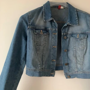 Blå croppad jeansjacka från H&M - Snygg blå croppad jeansjacka från H&M med knappar framtill och två bröstfickor med lock. Perfekt för en avslappnad stil. Jackan har en klassisk krage och långa ärmar.