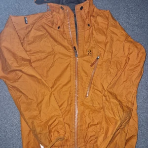 Orange vindjacka från Haglöfs - Säljer en orange vindjacka från Haglöfs med dragkedja och justerbar huva. Jackan har en bröstficka med dragkedja och elastiska ärmslut. Perfekt för blåsiga dagar och utomhusaktiviteter.