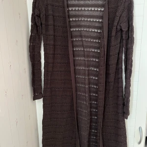 Brun stickad kofta från Capri Collection - Säljer en brun stickad kofta från Capri Collection i storlek M. Koftan har ett fint hålmönster och långa ärmar, perfekt för lager-på-lager-stil. Den är öppen framtill och ger en avslappnad look.