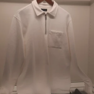 Vit långärmad tröja från Brand Island - Säljer en stilren vit långärmad tröja från Brand Island. Tröjan har en dragkedja framtill och en praktisk bröstficka. Perfekt för en avslappnad look.