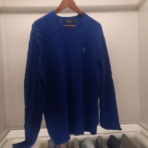 Blå stickad tröja från Polo Ralph Lauren - Säljer en snygg blå stickad tröja från Polo Ralph Lauren med det ikoniska gröna logotypbroderiet på bröstet. Tröjan har en klassisk kabelstickad design och långa ärmar, perfekt för kyligare dagar. Passar både till jeans och chinos för en stilren look.