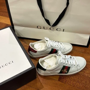 Vita sneakers från Gucci med broderad detalj - Snygga vita sneakers från Gucci med klassiska gröna och röda ränder samt en broderad insekt på sidan. Skorna har snörning och en stilren design som passar perfekt för en trendig look.