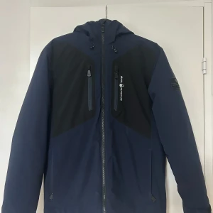 Blå jacka från Sail Racing - Säljer en blå jacka från Sail Racing med svarta detaljer och dragkedja framtill. Jackan har en sportig design med långärmade ärmar och en quiltad insida för extra värme. Perfekt för kyligare dagar. Den är på jätte god skick och är använd i 8 månader 