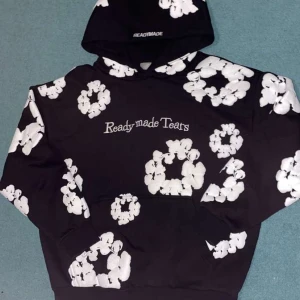 Svart hoodie med blommönster från Denim tears - Säljer en cool svart hoodie från Readymade med vita blommönster och texten 'Readymade Tears' på framsidan. Perfekt för en avslappnad stil med en unik touch. Hoodien har en klassisk passform och en bekväm huva.