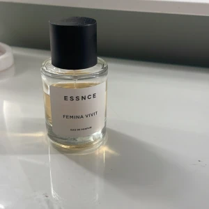 ESSNCE Femina Vivit Eau de Parfum - Elegant och sofistikerad parfym från ESSNCE med namnet Femina Vivit. Flaskan är stilren och genomskinlig med en svart kork och vit etikett. Perfekt för den som söker en unik doftupplevelse.