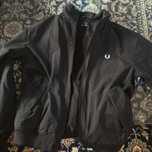 Svart bomberjacka från Fred Perry - Säljer en stilren svart bomberjacka från Fred Perry. Jackan har en dragkedja framtill och ribbade muddar vid ärmslut och nederkant. Den ikoniska loggan är broderad på bröstet. Perfekt för en cool och avslappnad look.