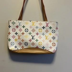 Vit axelväska med färgglatt mönster - Snygg vit axelväska med ett färgglatt monogrammönster. Väskan har en dragkedja upptill och bruna handtag. Insidan är fodrad med ett mörkt tyg med monogram. Perfekt för att ge en färgklick till din outfit!