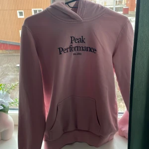 Rosa hoodie från Peak Performance - Säljer en snygg rosa hoodie från Peak Performance med broderad logga på bröstet. Den har en klassisk känguruficka och en bekväm huva. Perfekt för en avslappnad stil.