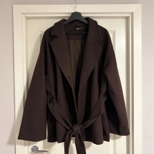 Brun kappa från gina tricot - Snygg brun kappa från gina tricot med knytband i midjan. Den har en klassisk design med långa ärmar och en stilren look. Perfekt för höst och vinterdagar när du vill hålla dig varm och trendig.
