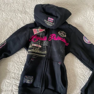 Mörkblå från Geographical Norway - Snygg mörkblå hoodie från Geographical Norway med  detaljer och tryck i rosa och beige. Den har en dragkedja framtill och en stor huva.  Aldrig använt