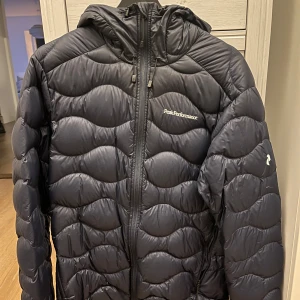 Mörkblå pufferjacka från Peak Performance - Säljer en mörkblå pufferjacka från Peak Performance med huva och dragkedja framtill. Jackan har ett quiltat mönster och är perfekt för kyligare dagar. Den är långärmad och har en stilren design.