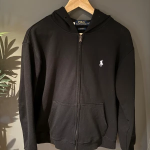 Svart hoodie från Polo Ralph Lauren - Säljer en stilren svart hoodie från Polo Ralph Lauren med dragkedja och det klassiska logotypbroderiet på bröstet. Tröjan har en bekväm passform och är perfekt för en avslappnad look. Den har långa ärmar och en praktisk huva.