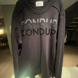Svart sweatshirt från Dondup - Säljer en svart sweatshirt från Dondup med tryckt logotyp i glänsande svart på framsidan. Tröjan har långa ärmar och en rund halsringning. Perfekt för en stilren och avslappnad look.