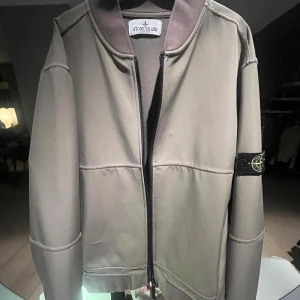 Beige jacka från Stone Island - Säljer en stilren beige jacka från Stone Island med dragkedja och den ikoniska loggan på ärmen. Jackan har en modern design med långa ärmar och en bekväm passform. Perfekt för dig som vill ha en snygg och funktionell jacka. Storlek 170 kids.