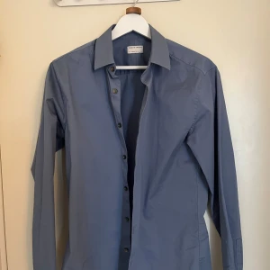 Blå skjorta från Tiger of Sweden - Snygg blå skjorta från Tiger of Sweden i extra slim passform. Skjortan har långa ärmar och knappar framtill. Perfekt för en stilren look.