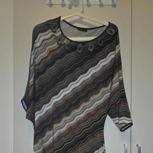 Mönstrad klänning från Binka - Säljer en unik klänning från Binka med vågigt mönster i brunt, svart och beige.har trekvartsärm och dekorativa detaljer vid halsringningen. Perfekt för en stilfull look.