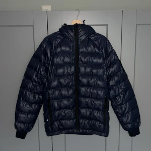 Mörkblå pufferjacka med huva - Säljer en snygg mörkblå pufferjacka från Peak Performance. Jackan är i stort sätt bra skick men vid den högra fickan finns det ett litet hål. Priset kan diskuteras vid snabb affär. Bara att höra av sig