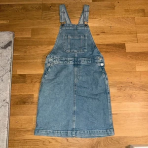 Jeansklänning med hängslen - Säljer en cool jeansklänning med justerbara hängslen och fickor fram och bak. Perfekt för en avslappnad look. Klänningen har knappar på sidorna för enkel på- och avtagning. Passar perfekt till sneakers eller sandaler!