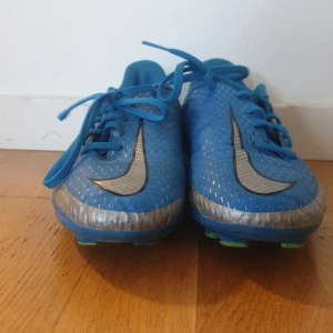 Hej - Snygga blå Nike fotbollsskor med silverdetaljer och snörning. Skorna har en sportig design med gröna dobbar och texten 'Phantom' på sidan. Perfekta för fotbollsplanen