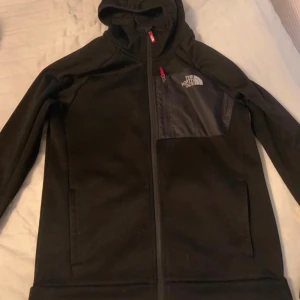 Svart fleecejacka från The North Face - Säljer en svart fleecejacka från The North Face med huva och dragkedja. Jackan har en bröstficka med dragkedja och logotyp på bröstet. Perfekt för kyliga dagar och outdoor-äventyr. 🏞️