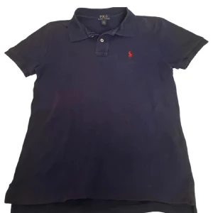 Mörkblå pikétröja från Ralph Lauren - Säljer en klassisk mörkblå pikétröja från Ralph Lauren med korta ärmar och en liten röd broderad logga på bröstet. Storlek 14-16 vilket motsvarar ungefär Xs.Tröjan har en krage och knappar framtill. Perfekt för en stilren look! 
