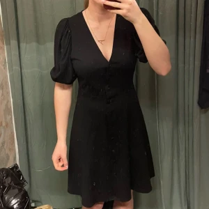 Svart klänning från H&M - Snygg svart klänning från H&M med v-ringning och knappar framtill. Den har korta puffärmar som ger en trendig look. Perfekt för olika tillfällen och lätt att matcha med accessoarer. Säljer pga att den aldrig är använd!🖤