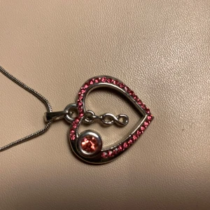 Halsband med hjärthänge och rosa stenar - Elegant halsband med ett hjärthänge dekorerat med små rosa stenar och en större rosa sten i mitten. Hänget har en unik design med en oändlighetssymbol inuti hjärtat. Kedjan är tunn och silverfärgad med justerbar längd. Använd 1 gång.