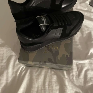 Svarta sneakers från Valentino - Snygga svarta sneakers från Valentino med klassisk snörning och stilren design. Skorna har en diskret camouflagemönstrad detalj och är tillverkade i skinn och syntetmaterial. Perfekta för en trendig look. Kvitto finns